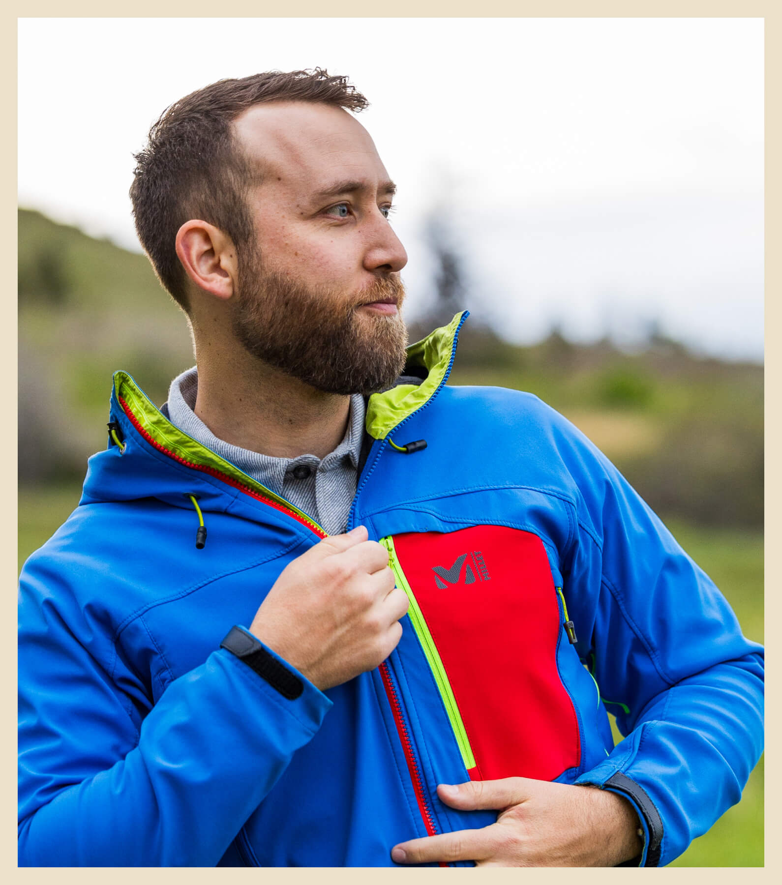 A bearded man wearing a blue and red jacket stands outdoors, looking to his right while pulling up the zipper on the jacket's chest pocket.