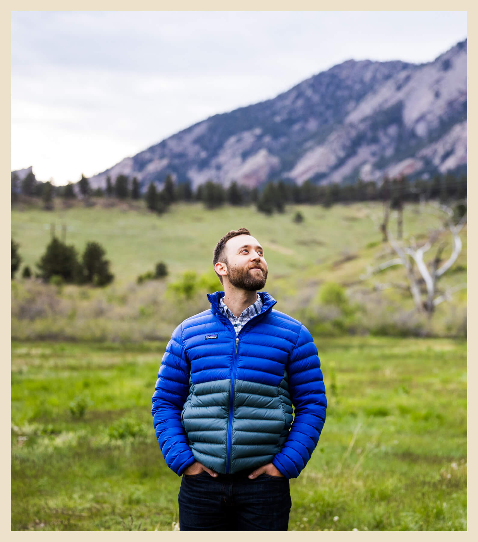 A person in a blue and gray jacket stands in a grassy field with mountains and a cloudy sky in the background.