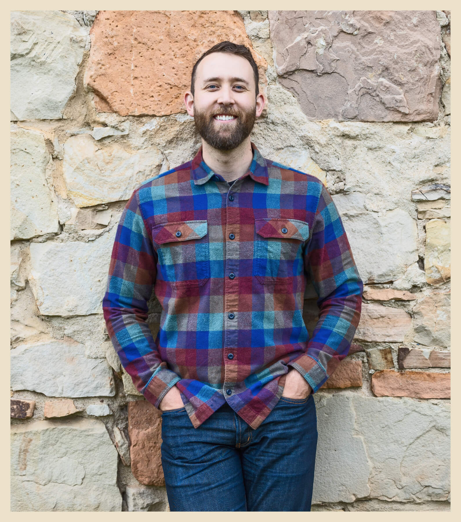 A man with a beard, wearing a plaid shirt and jeans, stands with his hands in his pockets against a rustic stone wall.