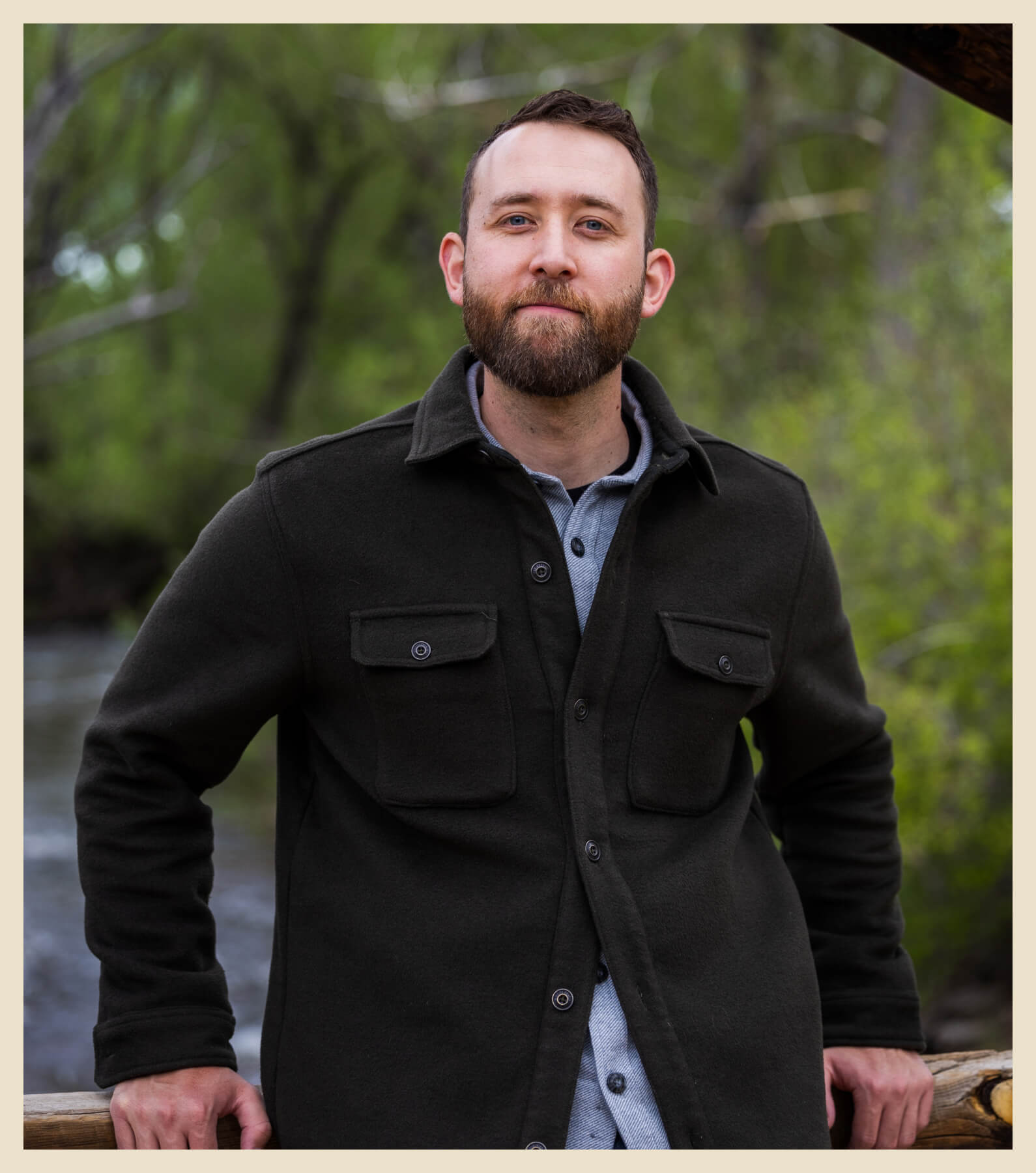 A man with a beard, wearing a dark jacket and shirt, stands outdoors in front of greenery and a body of water, leaning on a wooden railing.