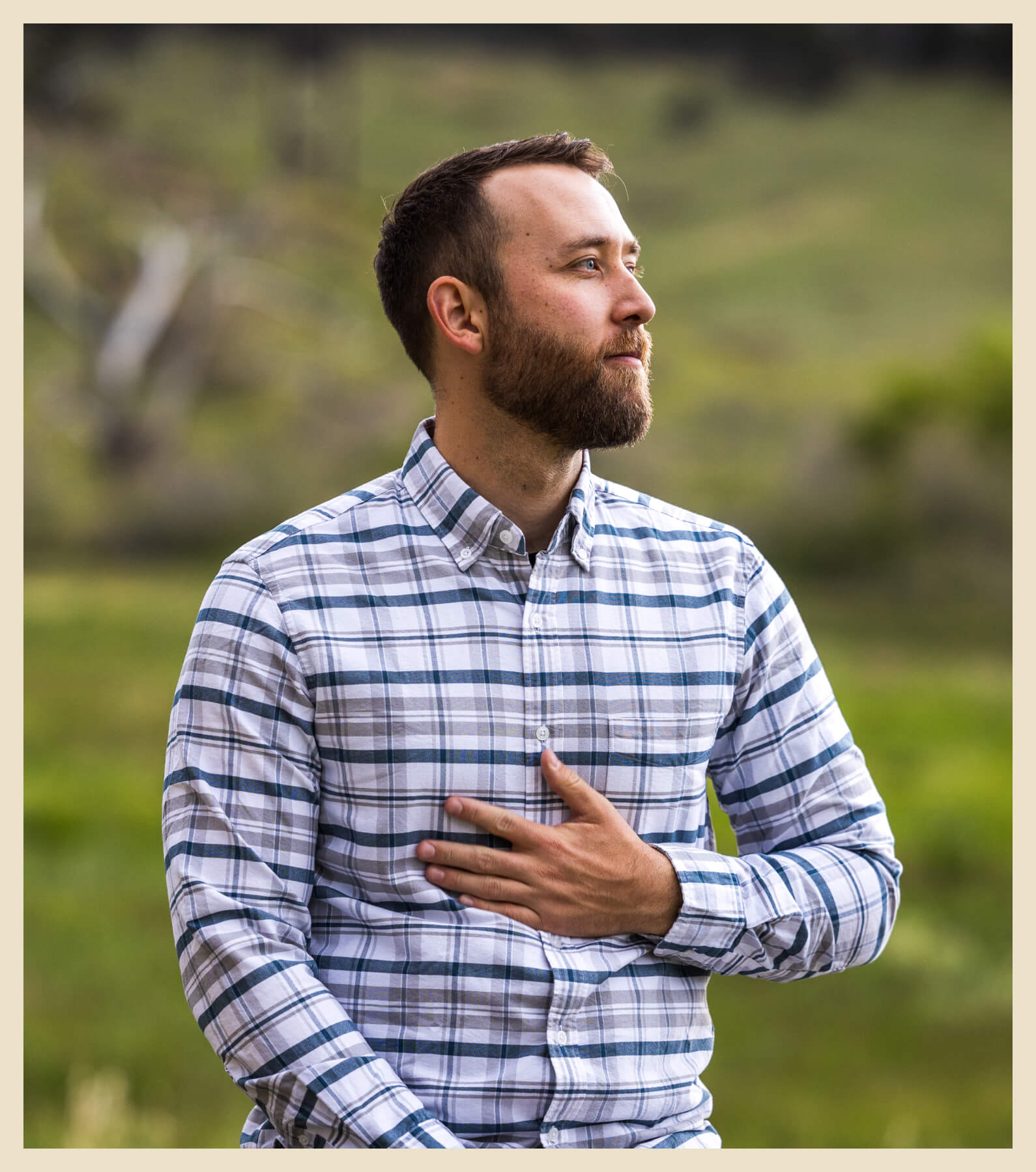 A man with a beard, wearing a plaid shirt, stands outdoors with one hand on his chest, looking to the side. Greenery and blurred natural background are visible.