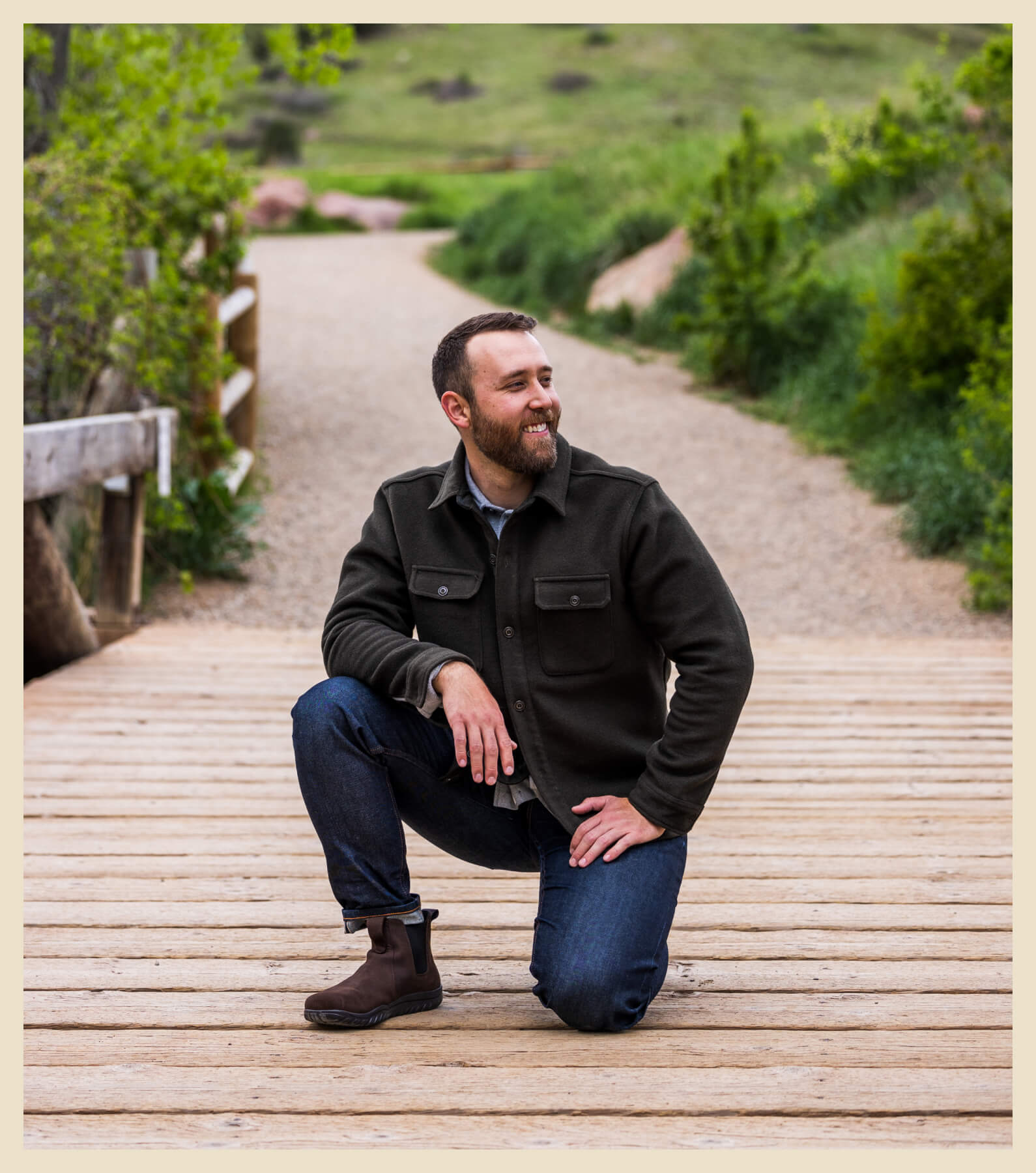 A man with a beard kneels on one knee on a wooden pathway in an outdoor setting with greenery in the background. He is wearing a dark jacket, jeans, and brown boots, looking slightly to his right.