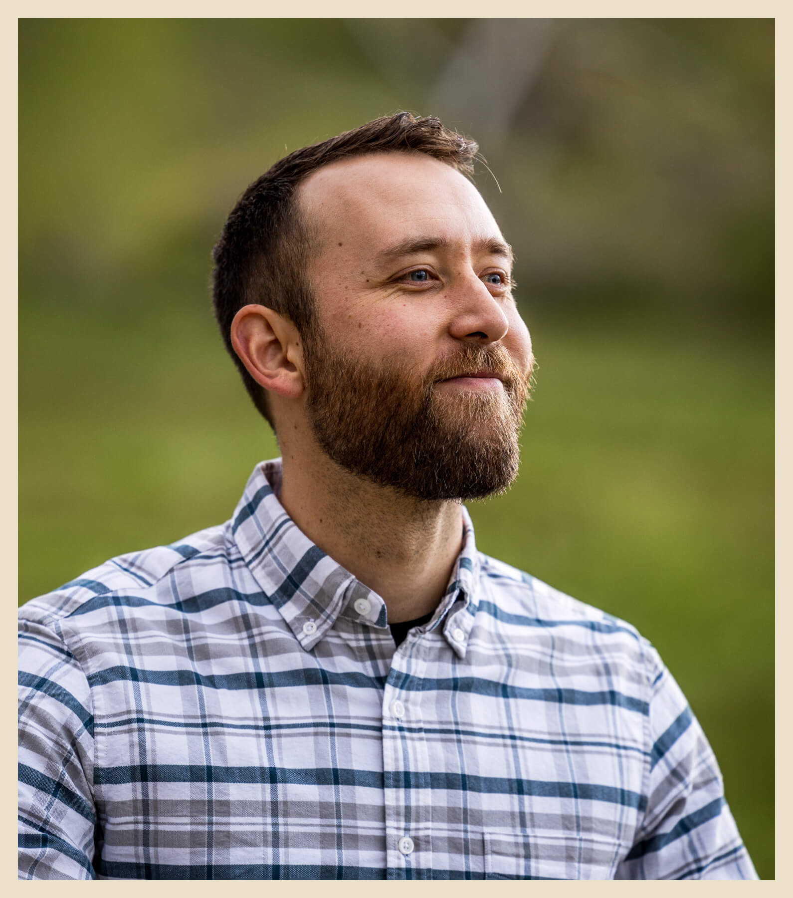 A bearded man in a plaid shirt stands outdoors, looking slightly upward with a neutral expression. The background is a blurred green landscape.