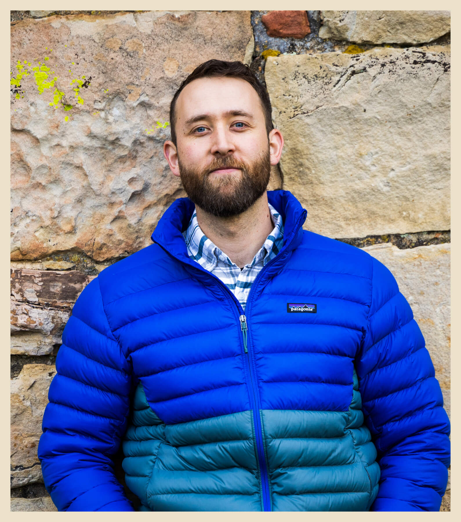 A man with a beard stands against a stone wall, wearing a blue and teal puffer jacket and a plaid shirt.